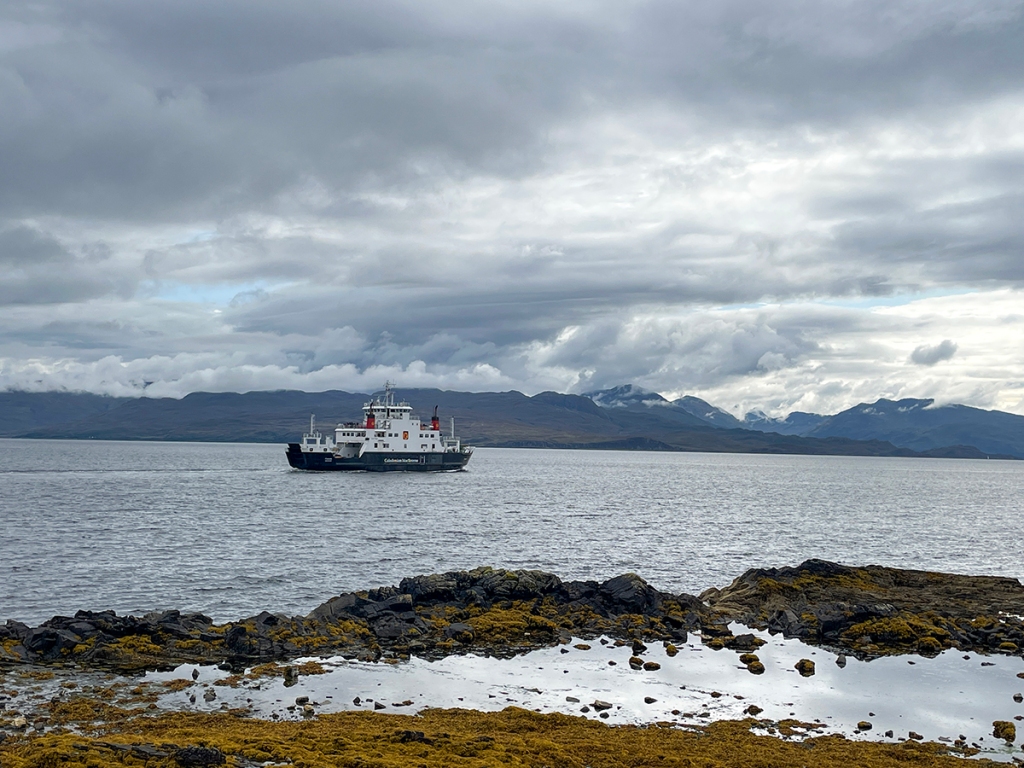A ferry crosses the water to the mainland, where you can see mountains and clouds.