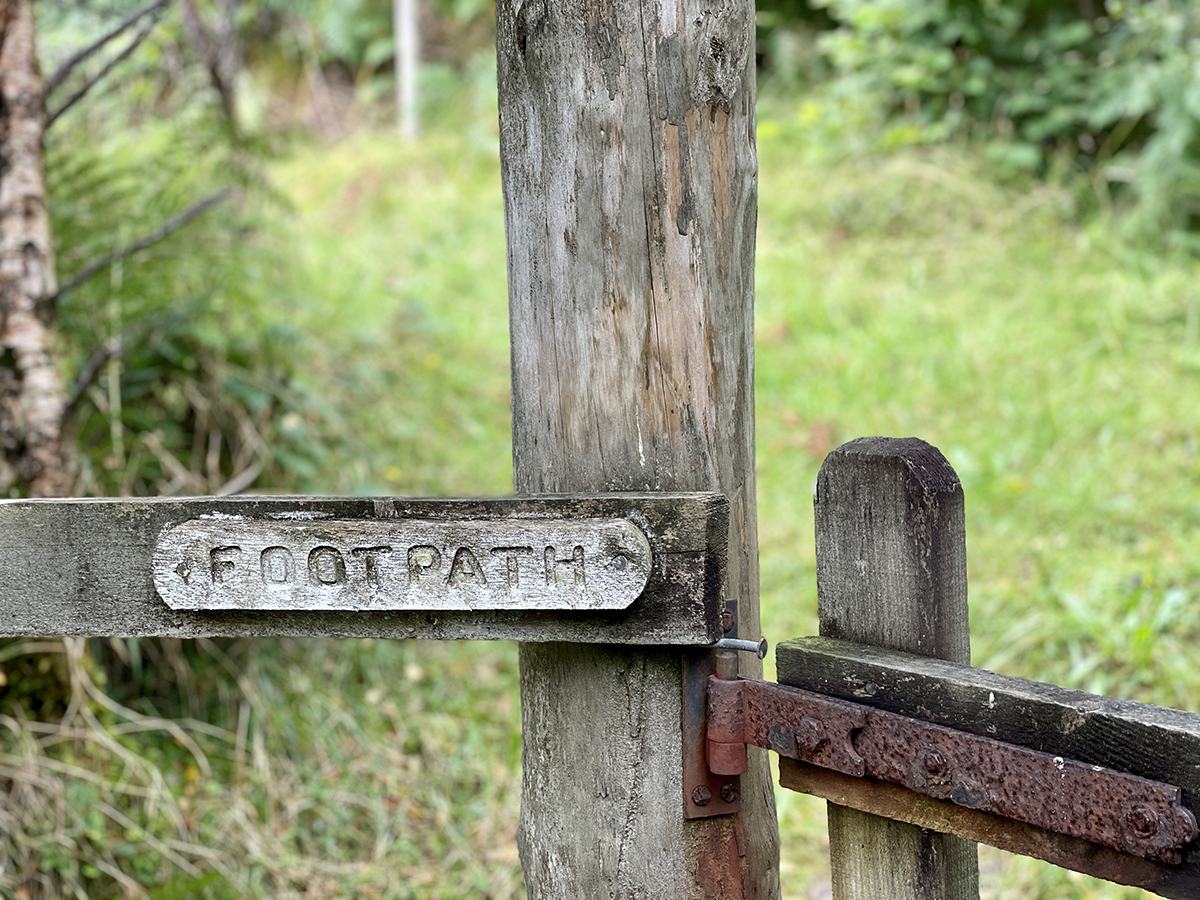 A wood post and rail fence with a rusty gate hinge and an engraved wooden sign that says FOOTPATH.