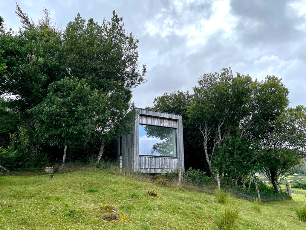 A wooden clad square building with a picture window looking out over a sheep field towards the Isle of Raasay, surrounded by trees.