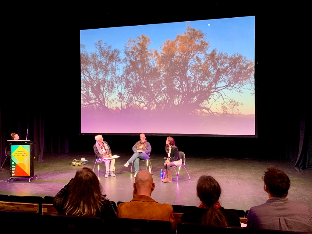 Photo of three people sitting on a stage with a video image behind them of trees at sunrise. The backs of four people in the audience can be seen in the foreground.