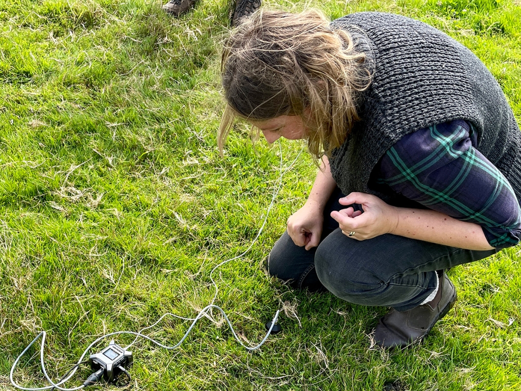 A woman is kneeling on the ground listening to the sound of the soil through earbuds connected to an audio recorder and geophone.