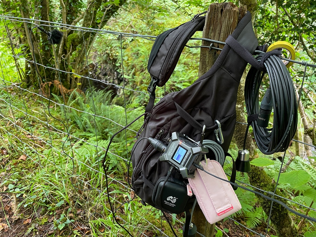 A black sling back with various audio recording equipment hanging from it, hanging on a ringlock fence post.